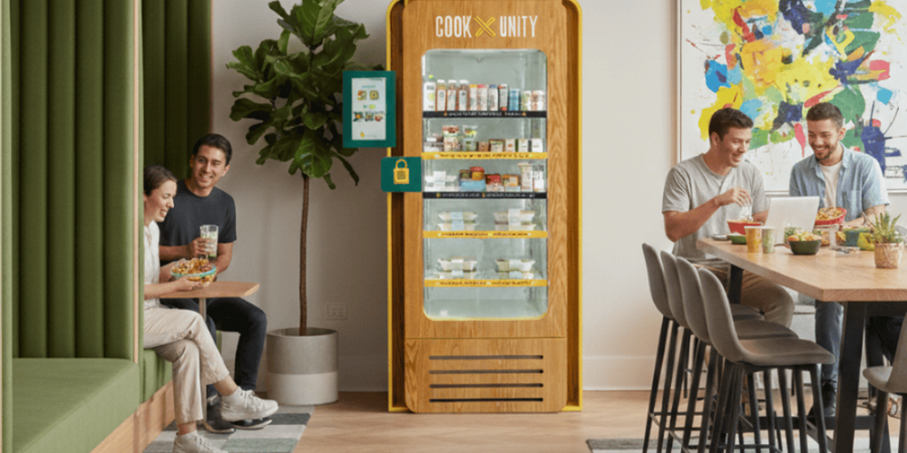 Two residents eating CookUnity meals next to a stocked CookUnity smart fridge in a bright modern apartment common area