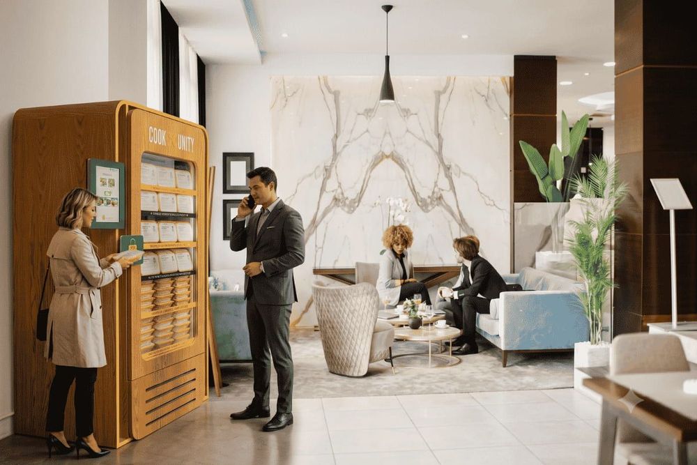 Resident using a smartphone to select a meal from a CookUnity smart fridge in an upscale apartment building lobby with marble walls and lounge seating