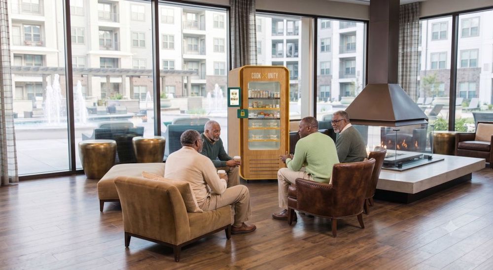 Four residents socializing in a luxury apartment building lounge next to a CookUnity smart fridge with no kitchen or staff required
