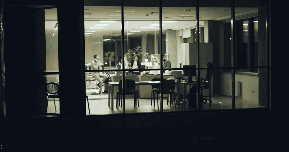 "Office workers visible through glass windows working late into the night at their desks, illustrating the food access challenge faced by night shift employees"